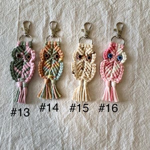 May include: Four handmade macrame owl keychains in various colors: green and pink, orange and green, cream, and pink. Each owl has a metal clasp and is labeled with a number: #13, #14, #15, and #16. The keychains are displayed on a textured, off-white surface.