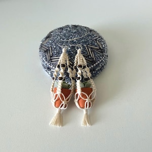 May include: Macrame earrings featuring small terracotta-colored pots with miniature succulents. The pots are suspended in cream-colored macrame holders, accented with dark brown beads and tassels. The earrings hang from silver-colored hooks.