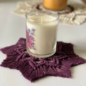 May include: A purple star-shaped crocheted coaster with gold accents sits on a white surface. A white candle with a floral design is sitting on top of the coaster.