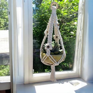 May include: A cream-colored macrame plant hanger with a wooden ring at the top, holding a green ceramic pot with a succulent plant. The macrame design includes decorative flower accents and a tassel at the bottom. The plant hanger is suspended in front of a window.