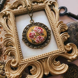 May include: A vintage-style pendant necklace featuring a round, embroidered floral design in a bronze-toned filigree setting. The pendant hangs from a delicate chain and is displayed within a gold ornate picture frame, evoking a sense of antique charm.