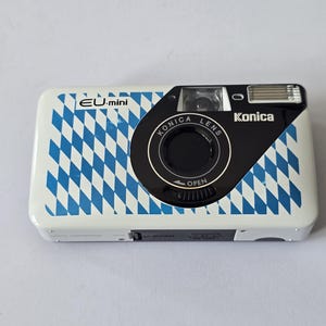 Op de afbeelding: Konica EU-mini camera met een wit en blauw patroon. De camera heeft een zwarte lens en flitser. Het ontwerp op de camera is een herhalend patroon van blauwe en witte ruiten. De woorden "Konica Lens" en "EU-mini" staan op de camera.