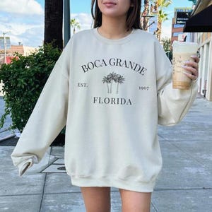 May include: A cream colored sweatshirt with a black graphic print of palm trees and the text "Boca Grande Est. 1907 Florida".