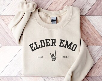 May include: A beige sweatshirt with the text "Elder Emo" in black, with "Est 1989" underneath. A black skeleton hand with a peace sign is between the text.