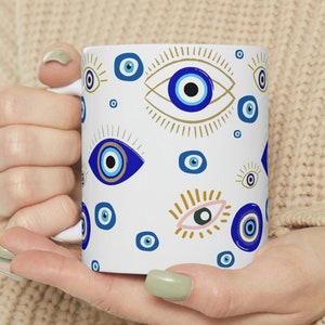May include: White ceramic mug featuring a blue and gold evil eye pattern. The mug is being held by a person's hands.