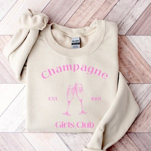 Op de afbeelding: Een beige sweatshirt met de tekst "Champagne Est 1991 Girls Club" in roze letters en een afbeelding van twee champagne glazen.