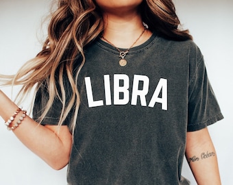 Può includere: Una donna indossa una maglietta grigia scura con la parola "LIBRA" stampata in bianco sul davanti. Indossa anche jeans blu e scarpe da ginnastica bianche.