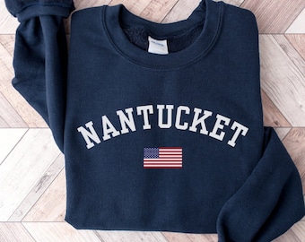 Könnte beinhalten: Marineblaues Sweatshirt mit dem weißen Wort "NANTUCKET" in Bogenform. Darunter befindet sich eine kleine amerikanische Flagge. Das Sweatshirt hat einen Rundhalsausschnitt und lange Ärmel mit gerippten Bündchen.