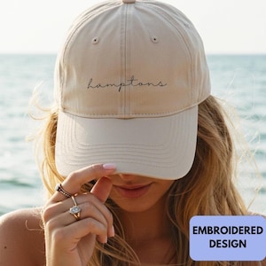 Könnte beinhalten: Eine beige Baseballkappe mit dem Wort hamptons, das in einer dunklen, dünnen Schreibschrift auf die Vorderseite gestickt ist. Die Kappe hat eine strukturierte Krone, einen gebogenen Schirm und einen kleinen Knopf oben auf dem Stoff.