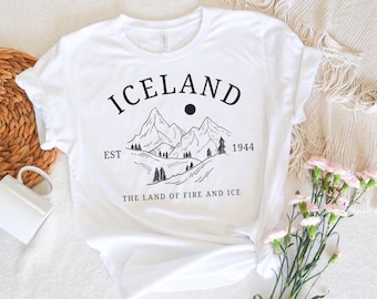 Puede incluir: Camiseta blanca con un diseño gráfico negro de montañas y un sol. El texto "ICELAND" está por encima de las montañas, y el texto "EST 1944 THE LAND OF FIRE AND ICE" está por debajo de las montañas.