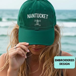 Può includere: Un cappellino da baseball verde scuro con la scritta "NANTUCKET" ricamata in bianco, insieme a un disegno di un faro. Il cappellino è indossato da una persona. Le parole "EMBROIDERED DESIGN" sono in un riquadro azzurro.