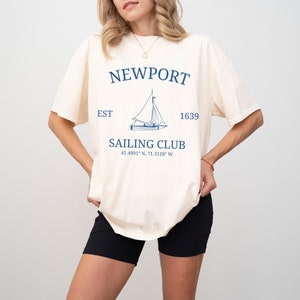 Op de afbeelding: Een crèmekleurig T-shirt met een blauwe zeilbootgrafiek en de tekst "NEWPORT SAILING CLUB EST 1639 41.4901° N, 71.3128° W"