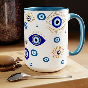 May include: White ceramic mug with a blue rim and handle. The mug features a repeating pattern of blue evil eyes with gold accents on a white background.