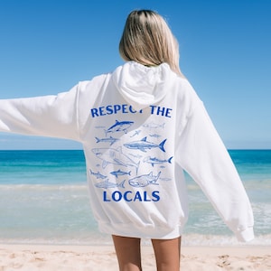 Könnte beinhalten: Weißer Hoodie mit einem blauen Aufdruck von Haien und dem Text "Respect the Locals" auf der Rückseite. Der Hoodie wird von einer Person getragen, die an einem Strand steht.