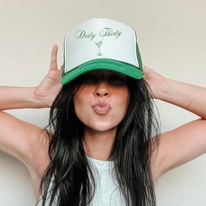 May include: White and green trucker hat with the text "Dirty Thirty" in green script, along with a martini glass graphic. The hat has a green brim and mesh back.