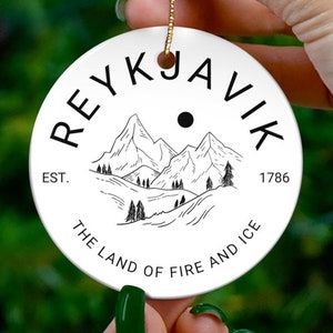 Könnte beinhalten: Weißer Keramik-Weihnachtsbaumschmuck mit einer Schwarzweißillustration von Bergen und dem Text "REYKJAVIK EST. 1786 THE LAND OF FIRE AND ICE".