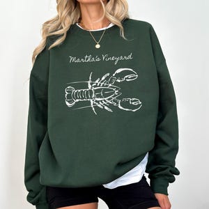 May include: Dark green sweatshirt with the text "Martha's Vineyard" and a white lobster illustration. The sweatshirt has a white crew neck and long sleeves. The model is wearing black shorts.
