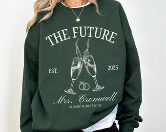 Puede incluir: Una sudadera verde oscuro con el texto "THE FUTURE" en blanco, con un gráfico de dos copas de champán brindando, y el texto "EST. 2025" y "Mrs. Cromwell 44.3386° N, 68.2733° W".