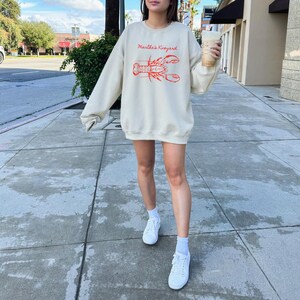 May include: Cream-colored sweatshirt with the text "Martha's Vineyard" above a red lobster illustration. The long-sleeved top is worn with white sneakers and socks. The person is holding a beverage in a clear cup.