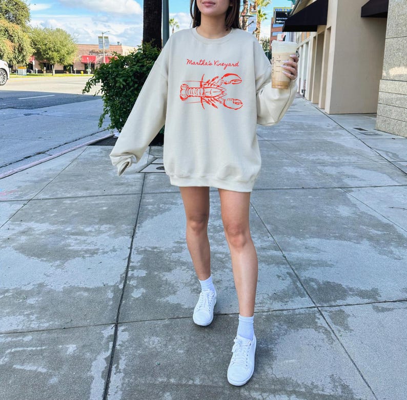 May include: Cream-colored sweatshirt with the text "Martha's Vineyard" above a red lobster illustration. The long-sleeved top is worn with white sneakers and socks. The person is holding a beverage in a clear cup.