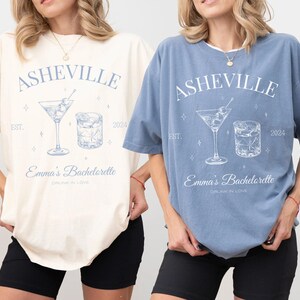 May include: Two women wearing oversized t-shirts with a blue graphic design. The shirts say "Asheville Est. 2024" and feature a martini glass and a rocks glass. The text below the graphic says "Emma's Bachelorette Drunk in Love". One shirt is ivory and the other is blue jean.