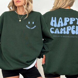 Peut inclure: Sweat-shirt vert foncé avec l'inscription "HAPPY CAMPER" en bleu clair au dos et un smiley avec les mots "HAPPY CAMPER" sur le devant. Le texte "Ashley's Bachelorette" est en dessous du texte au dos.
