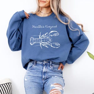 May include: A blue sweatshirt with the words "Martha's Vineyard" above a white lobster graphic. The sweatshirt is paired with blue jeans. The overall theme is coastal and nautical.