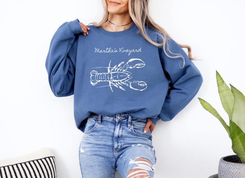 May include: A blue sweatshirt with the words "Martha's Vineyard" above a white lobster graphic. The sweatshirt is paired with blue jeans. The overall theme is coastal and nautical.