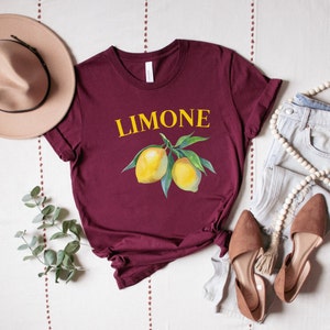 May include: A maroon t-shirt with the word "LIMONE" in yellow above a watercolor illustration of lemons on a branch. The shirt is styled with a beige hat, jeans, and brown shoes.