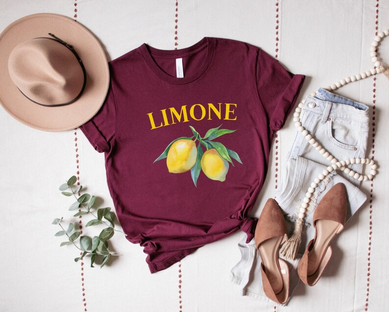 May include: A maroon t-shirt with the word "LIMONE" in yellow above a watercolor illustration of lemons on a branch. The shirt is styled with a beige hat, jeans, and brown shoes.