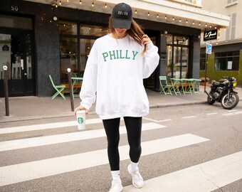 May include: A white sweatshirt with the word "PHILLY" in green lettering. The person is wearing black leggings and a black baseball cap with the number "11:11" on it.