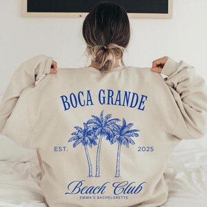 May include: A beige sweatshirt with a blue graphic design that says "Boca Grande Beach Club" with palm trees and the text "Est. 2025" and "Emma's Bachelorette"