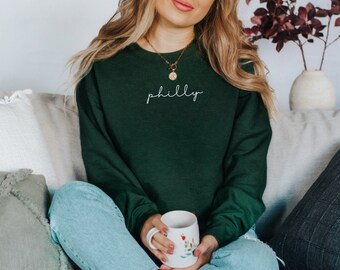 May include: A dark green sweatshirt with the word "philly" in white script. The sweatshirt is long-sleeved and has a crew neck. The person is wearing blue jeans and holding a white mug with a floral design.