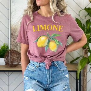 May include: Mauve-colored t-shirt with the word "LIMONE" in yellow above a watercolor illustration of two lemons and green leaves. The shirt is knotted at the waist and paired with blue denim shorts.