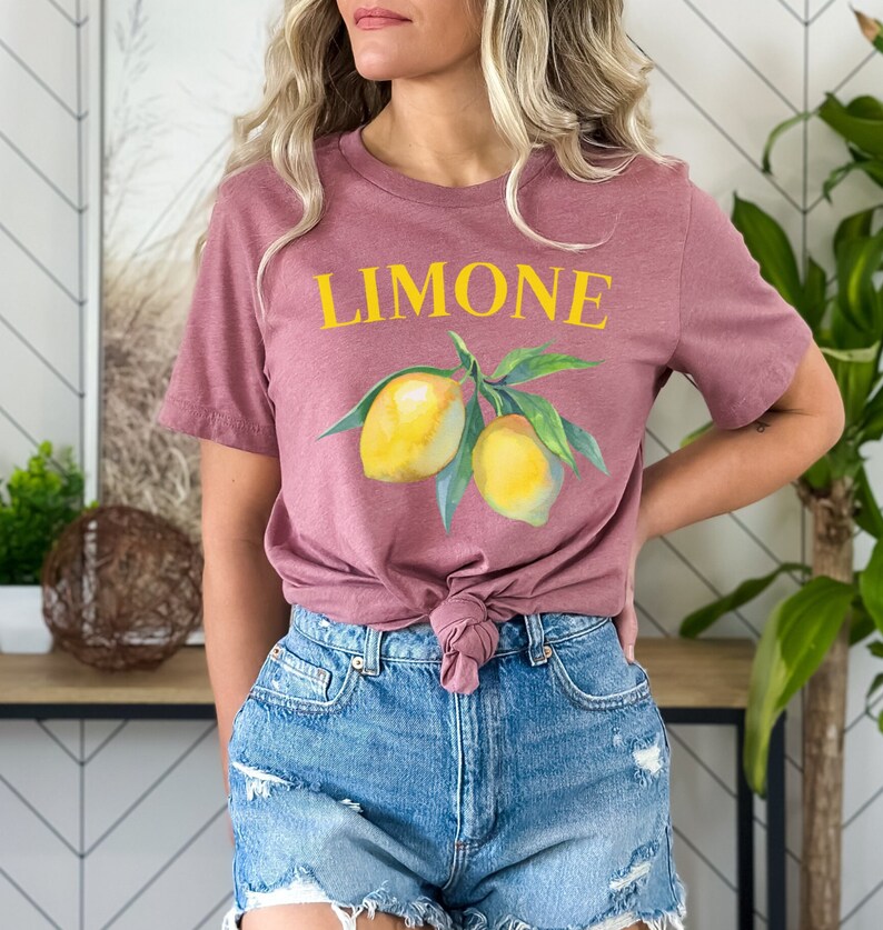 May include: Mauve-colored t-shirt with the word "LIMONE" in yellow above a watercolor illustration of two lemons and green leaves. The shirt is knotted at the waist and paired with blue denim shorts.
