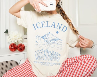 Op de afbeelding: Een wit T-shirt met een blauwe afbeelding van een bergketen en de tekst "IJsland, Het Land van Vuur en IJs, Gevestigd in 1944".