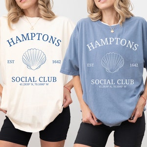Könnte beinhalten: Zwei übergroße T-Shirts mit einem grafischen Design mit dem Text "HAMPTONS SOCIAL CLUB" mit einer Muschel und dem Text "EST 1642" und Koordinaten. Das T-Shirt links ist elfenbeinfarben und das T-Shirt rechts ist jeansblau.