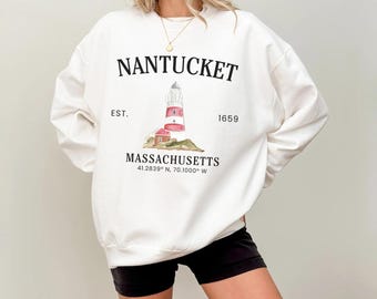 Könnte beinhalten: Weißes Sweatshirt mit dem Wort "NANTUCKET" in Schwarz, über einem Aquarellbild eines rot-weißen Leuchtturms. Unter dem Leuchtturm stehen "MASSACHUSETTS" und Koordinaten. Auf dem Sweatshirt steht auch "EST. 1659".