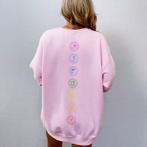 May include: Pink sweatshirt with a vertical row of seven colorful chakra symbols printed on the back. The symbols are in a circle with a decorative border.