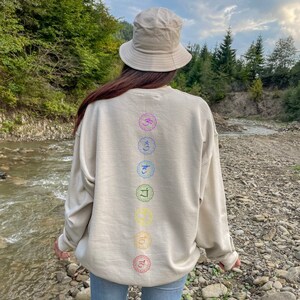 May include: A beige sweatshirt with a vertical row of seven colorful chakra symbols printed on the back. The symbols are in a circle with a flower design. The sweatshirt is worn by a person wearing a beige bucket hat and blue jeans. The person is standing in a forest setting near a stream.