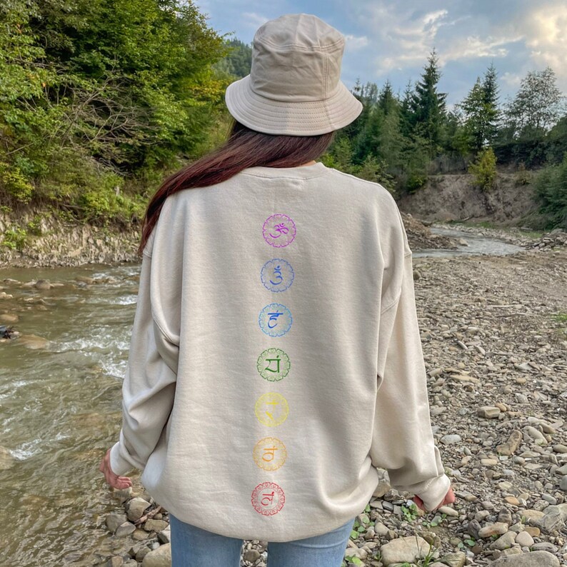 May include: A beige sweatshirt with a vertical row of seven colorful chakra symbols printed on the back. The symbols are in a circle with a flower design. The sweatshirt is worn by a person wearing a beige bucket hat and blue jeans. The person is standing in a forest setting near a stream.