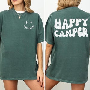 Peut inclure: Un t-shirt vert avec un smiley blanc et le texte "Happy Camper" sur le devant et le texte "Happy Camper" en police ondulée au dos.