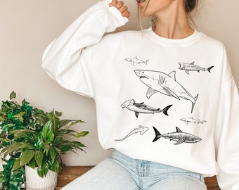 May include: White sweatshirt with a black and white illustration of different shark species. The design includes a great white shark, hammerhead shark, and other shark types. A casual, comfortable top for ocean enthusiasts.