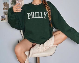 May include: A green sweatshirt with the word "PHILLY" printed in white on the front. The sweatshirt is being worn by a person with long brown hair. The person is wearing white cowboy boots.