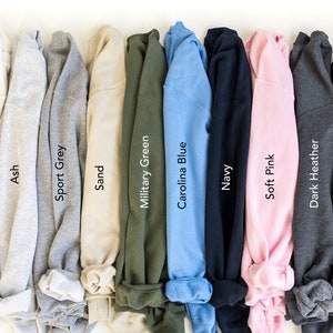 May include: A selection of ten folded sweatshirts in various colors, including white, ash, sport grey, sand, military green, carolina blue, navy, soft pink, dark heather, and black.