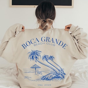 May include: A beige sweatshirt with a blue graphic print of palm trees and the text "Boca Grande Beach Club" and "May, 2025".