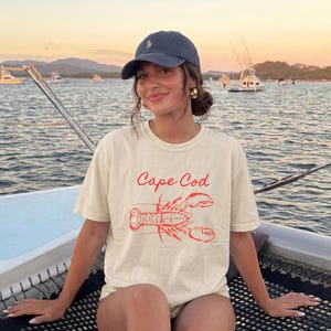 Puede incluir: Una gorra de béisbol azul marino con un pequeño logotipo bordado y una camiseta de manga corta color crema con un gráfico rojo de una langosta y el texto Cape Cod impreso en letra cursiva en el centro de la zona del pecho.