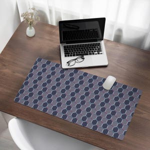Op de afbeelding: Een grote, donkergrijze en paarse muismat met een zeshoekig patroon op een houten bureau met een laptop en een bril erop.