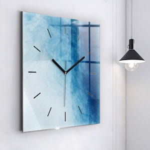 Op de afbeelding: Een vierkante wandklok met een glad, glanzend oppervlak en een abstracte aquarelprint in tinten licht- en donkerblauw. De wijzerplaat is voorzien van dunne, zwarte streepjes als uurmarkeringen en eenvoudige zwarte uur-, minuut- en secondewijzers.