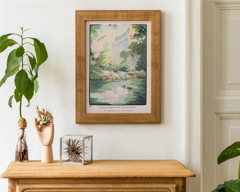 May include: A framed vintage illustration of the Hotel Plaza in New York City, seen from Central Park. The illustration is in a wooden frame and features a view of the hotel with swans in the foreground.